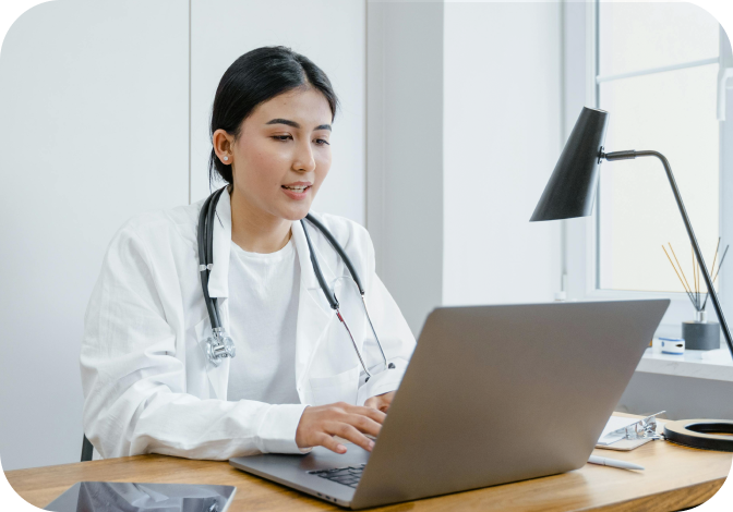 A doctor sitting at a wooden table reviewing notes on her computer.