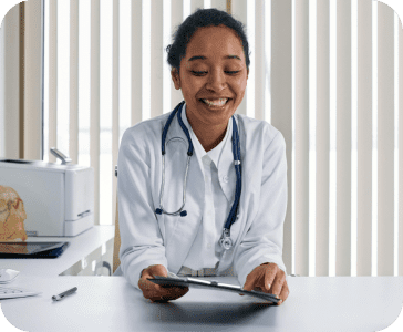 A doctor smiling while reading a medical chart. She's sitting down with a white lab coat on and a blue stethiscope